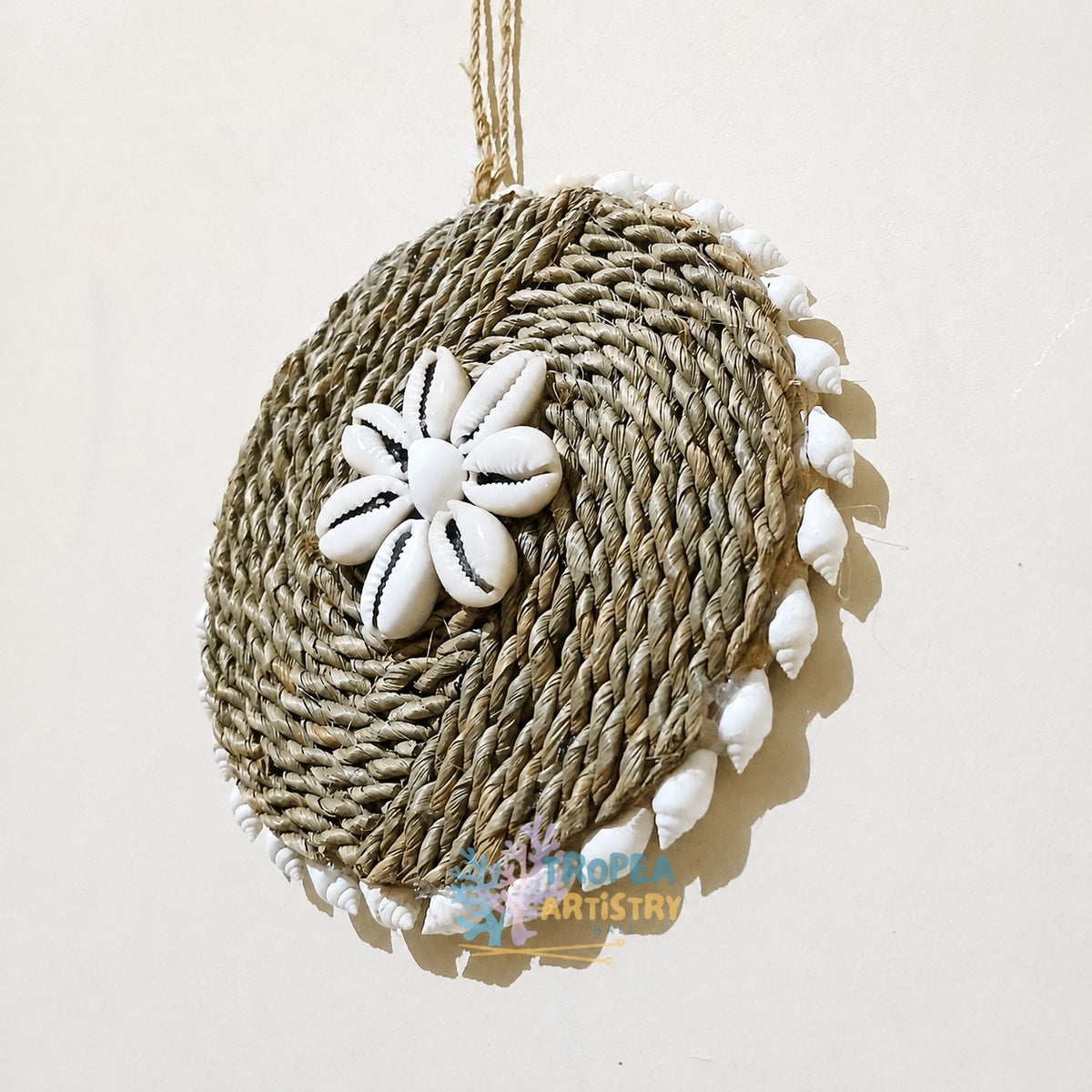 A round woven raffia wall hanging ornament decorated with a white cowrie shell flower in the center and white cowrie shells trimmed around the edge, hanging against a plain off-white wall.