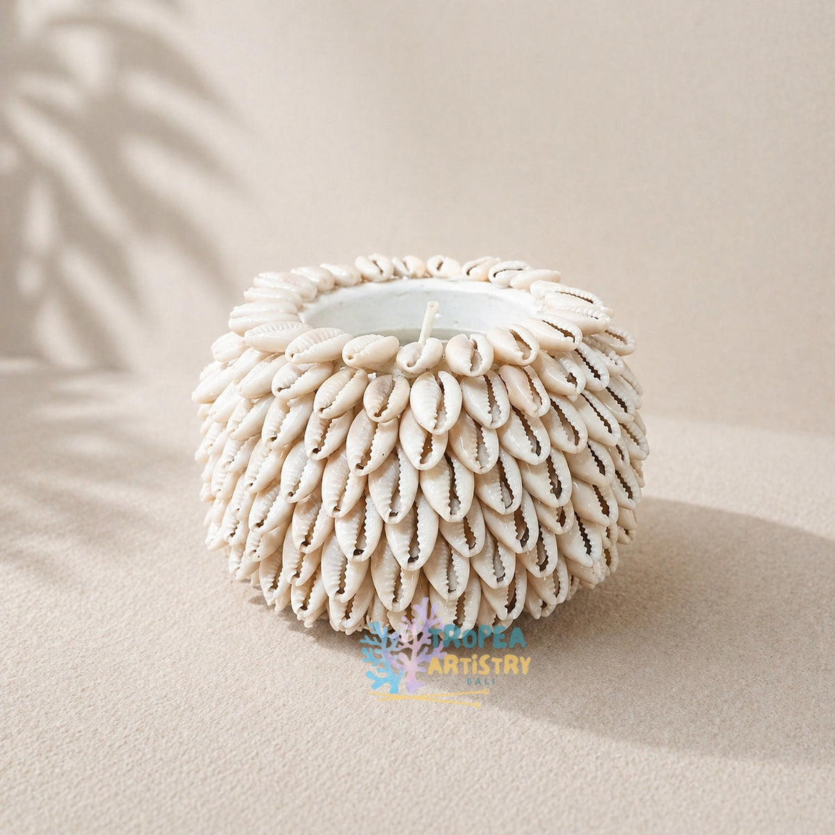 A round white candle holder decorated with numerous small cowrie shells, set on a beige textured surface with soft palm leaf shadows.