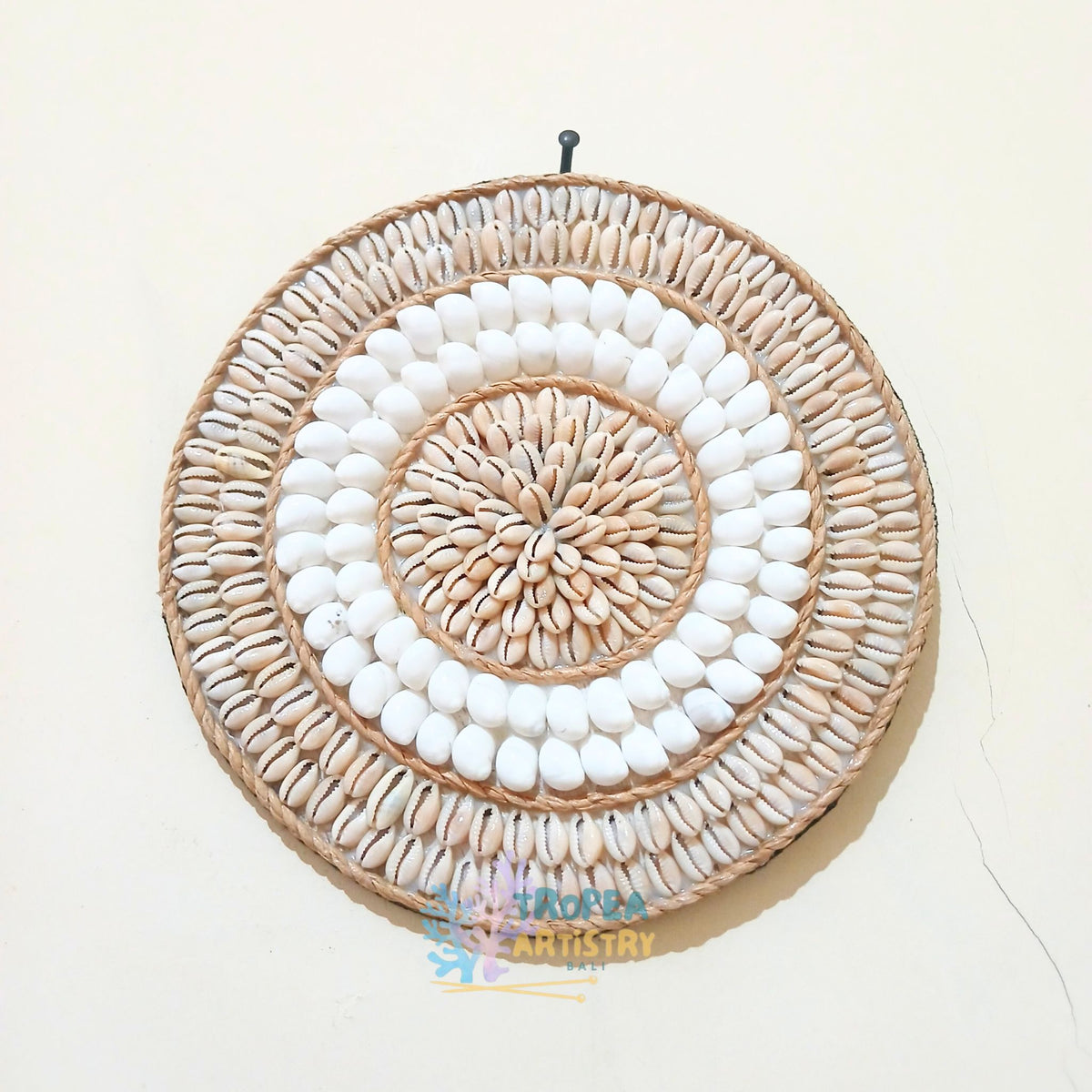 A circular woven wall hanging made from natural tan raffia and white seashells, arranged in concentric rings, shown hanging on a plain off-white wall.