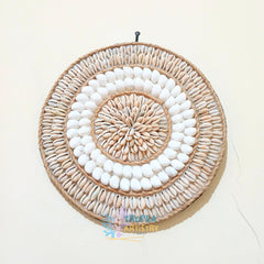 A circular woven wall hanging made from natural tan raffia and white seashells, arranged in concentric rings, shown hanging on a plain off-white wall.