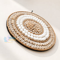 An oval woven wall hanging made from natural raffia rope and rows of white cowrie shells, photographed on a clean off-white background with a Tropen Artistry Bali watermark in the lower left.