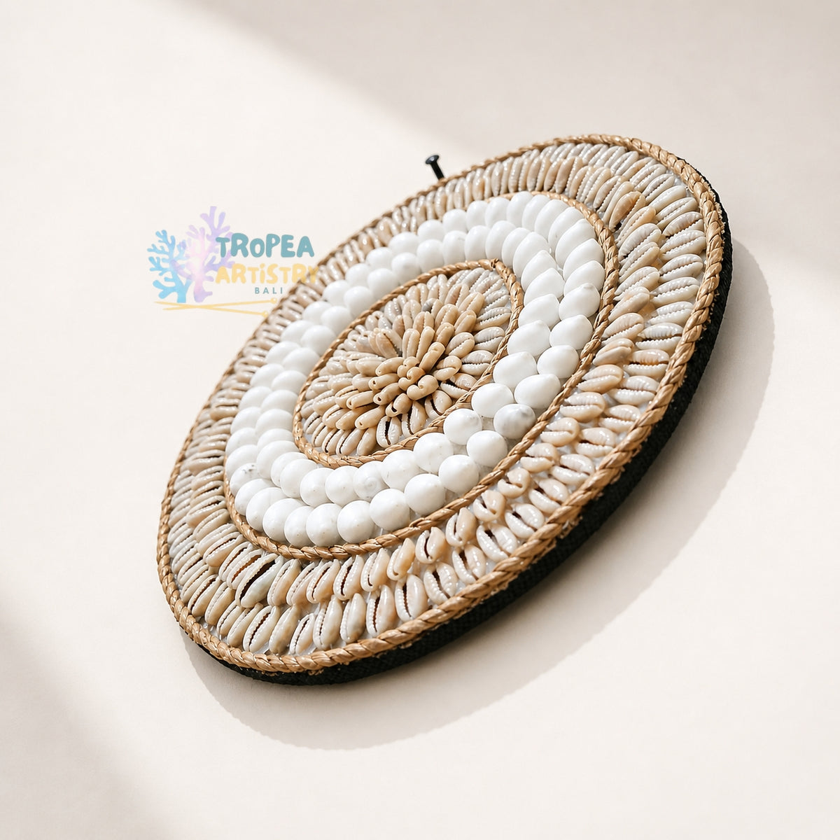A circular woven wall hanging made from natural fiber and white shell beads, photographed against a warm off-white background with a visible shadow. A Tropea Artisan watermark appears on the left side.