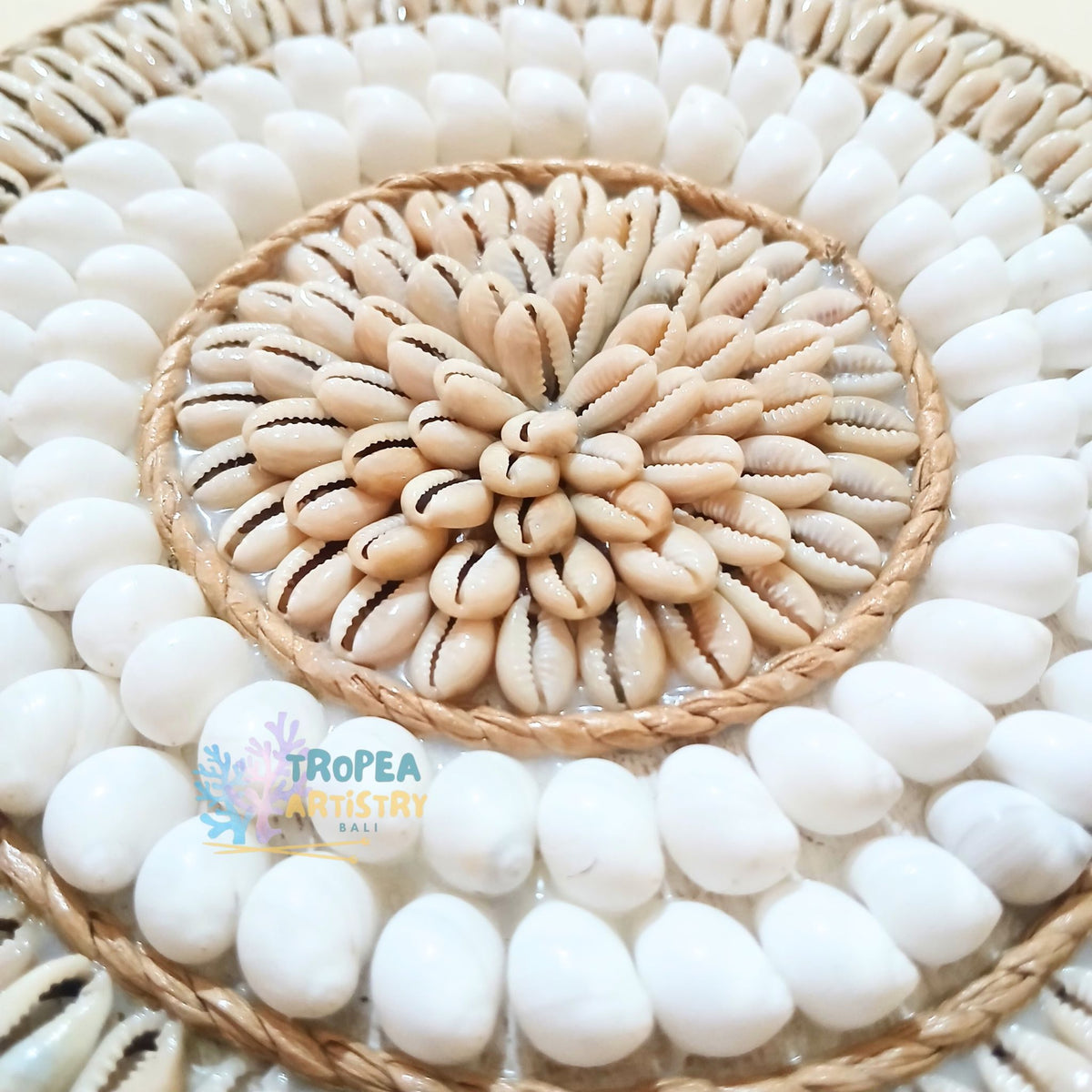 A circular decorative wall art piece made from white cowrie shells and natural beige shell curls arranged in concentric circles on a woven base, with a visible Tropea Artistry logo watermark near the bottom left.