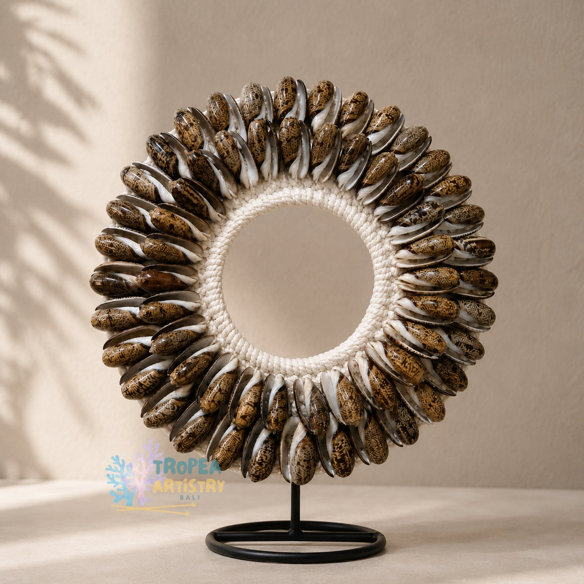 A circular decorative wall piece made of many small brown and cream seashell-like pieces arranged around a white rope center, mounted on a black metal stand against a warm beige wall with soft shadowing.