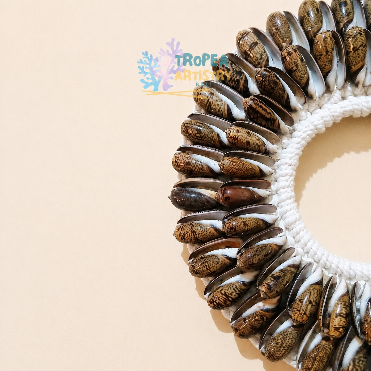 A circular wall hanging made from many small seashell halves arranged around a white braided ring, photographed on a plain warm beige background. A TroPia Artistry watermark appears near the top center.