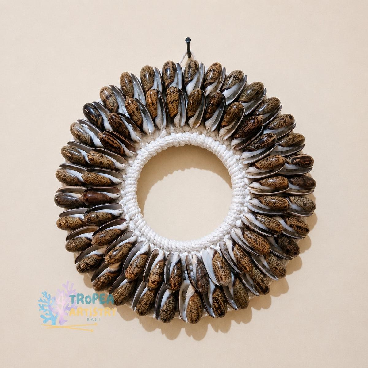 A circular seashell wreath wall hanging with a white braided rope center, photographed against a plain beige wall. A small black hook is attached at the top, and a Tropfe Artisan Klu watermark appears in the lower-left corner.