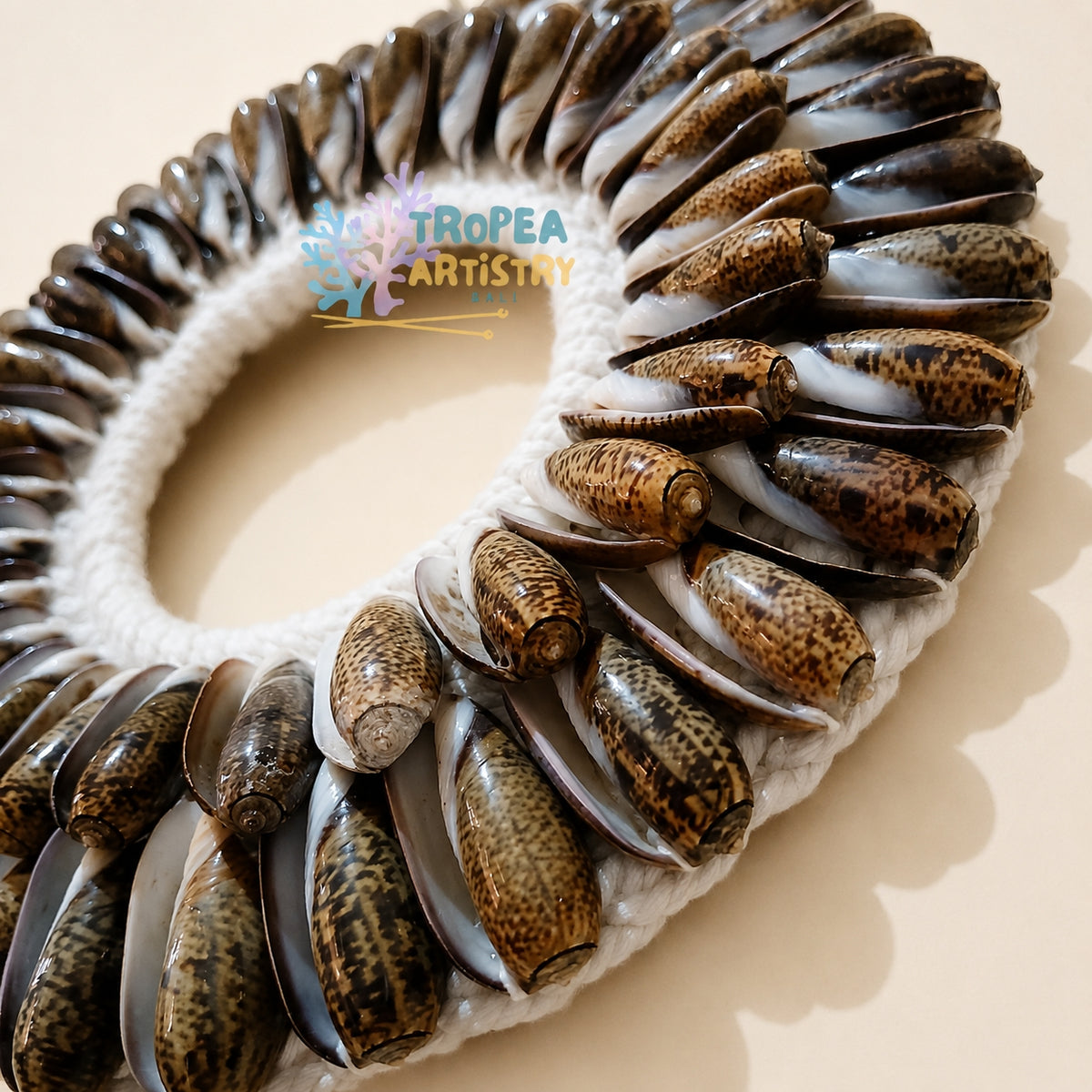 A circular handmade wall art piece featuring a white crochet border with numerous brown and black patterned cowrie shells arranged around the edge, photographed on a warm beige background. A 'TROPEA ARTISTRY' watermark is visible in the center.