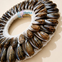 A circular handmade wall art piece featuring a white crochet border with numerous brown and black patterned cowrie shells arranged around the edge, photographed on a warm beige background. A 'TROPEA ARTISTRY' watermark is visible in the center.