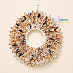 A circular decorative wall hanging made of many pointed, fan-like elements in metallic silver, warm orange, and black, arranged around a white rope center on a plain beige wall. A Tropea Artistry Bali watermark appears on the right side.