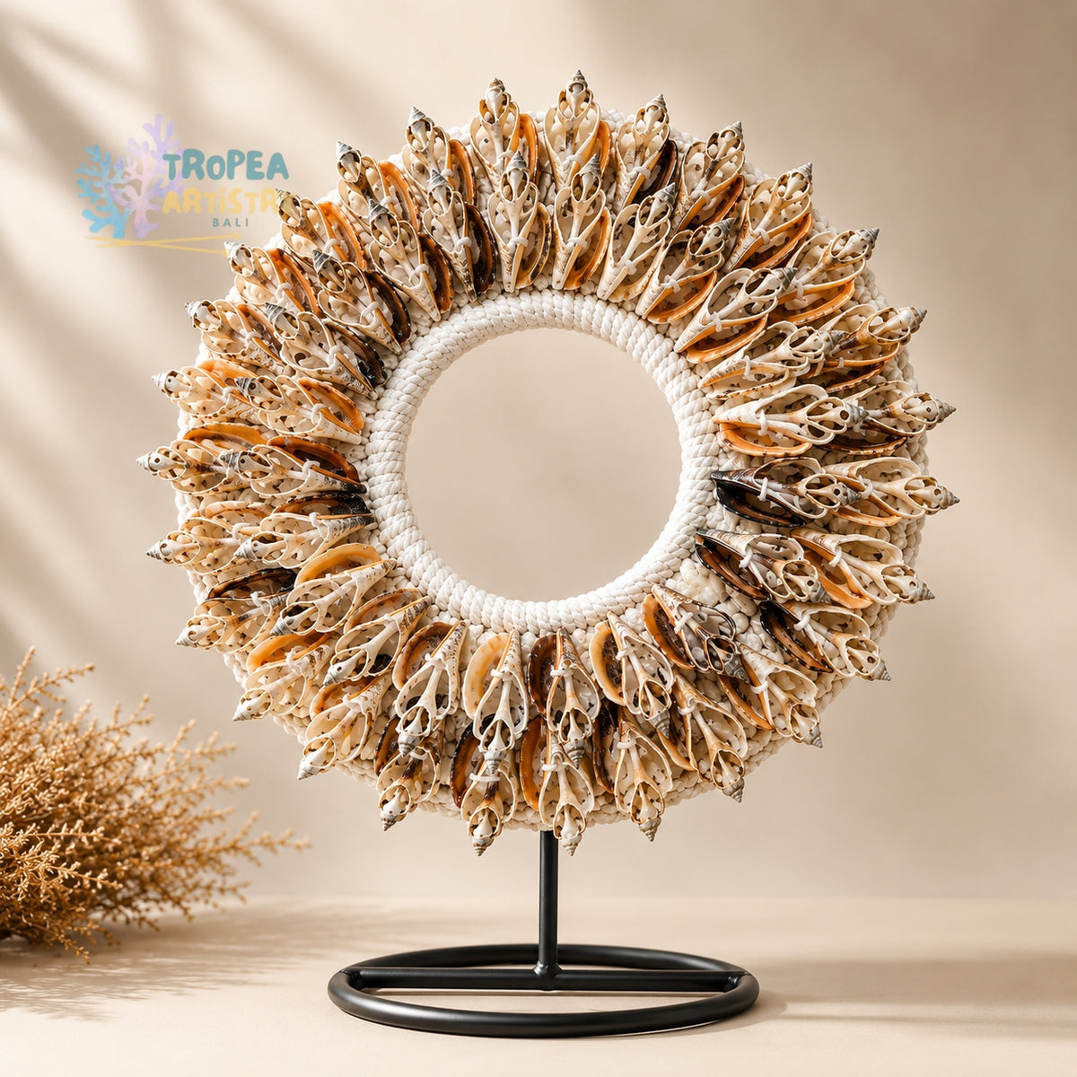 A circular decorative wreath made of many dried seashell-like pieces in beige, brown, and white tones, mounted on a white rope ring and displayed on a black stand against a warm beige background. A small Astropea logo appears in the upper left, and a dried plant prop sits at lower left.