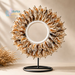 A circular decorative wreath made of many dried seashell-like pieces in beige, brown, and white tones, mounted on a white rope ring and displayed on a black stand against a warm beige background. A small Astropea logo appears in the upper left, and a dried plant prop sits at lower left.