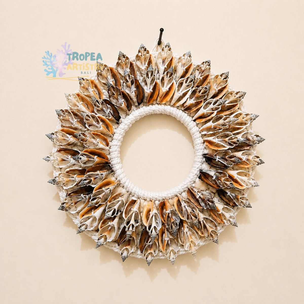 A circular shell wall hanging wreath made of numerous small seashell fragments arranged around a white rope center, shown against a plain beige background. A Tropea Artisan logo is visible in the upper left.