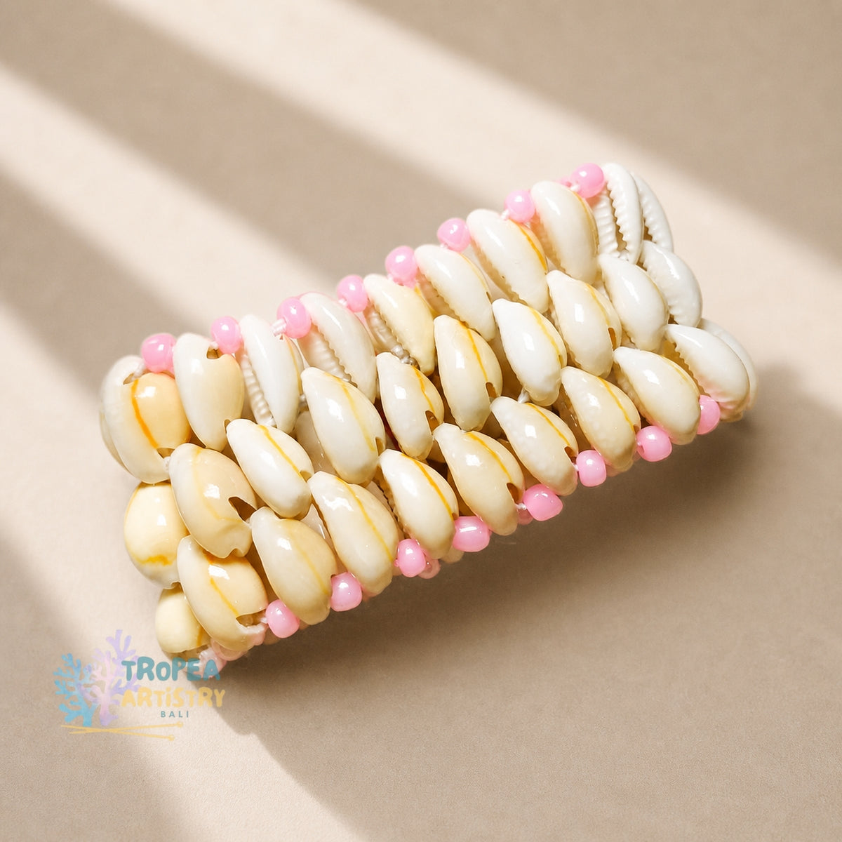 A handmade cuff bracelet made of cream-colored cowrie shells with small pink beads, displayed on a neutral beige background with soft diagonal shadows.