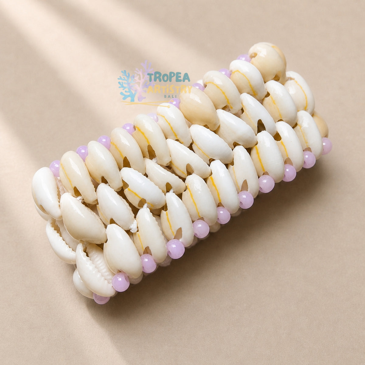A woven bracelet made of cream and white cowrie shells, accented with small light purple beads, displayed on a neutral beige background.