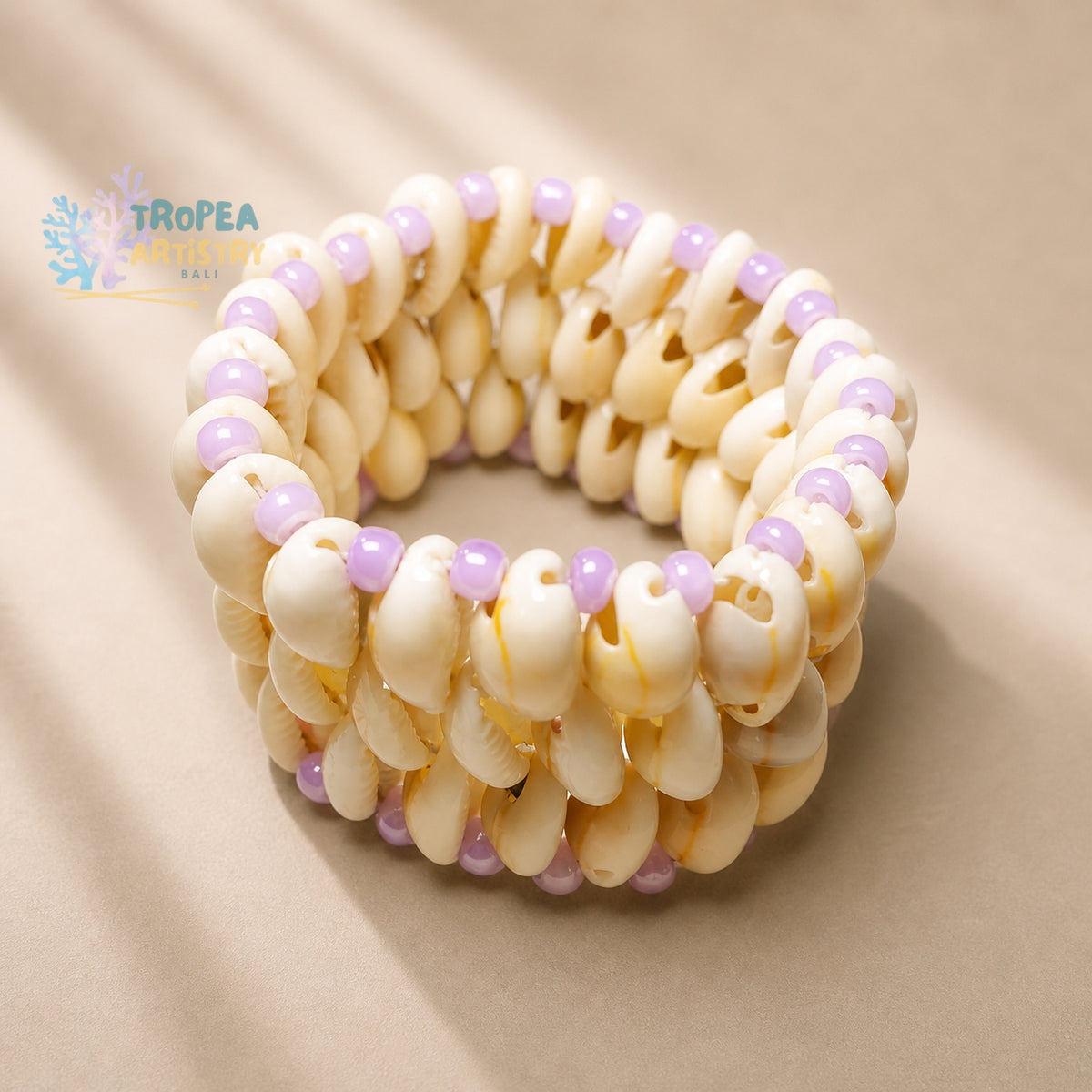 A beaded stretch bracelet made of cream-colored cowrie shells with small lavender purple beads, displayed on a neutral beige background. A 'TROPEA BAILI' brand logo is visible in the upper left corner.