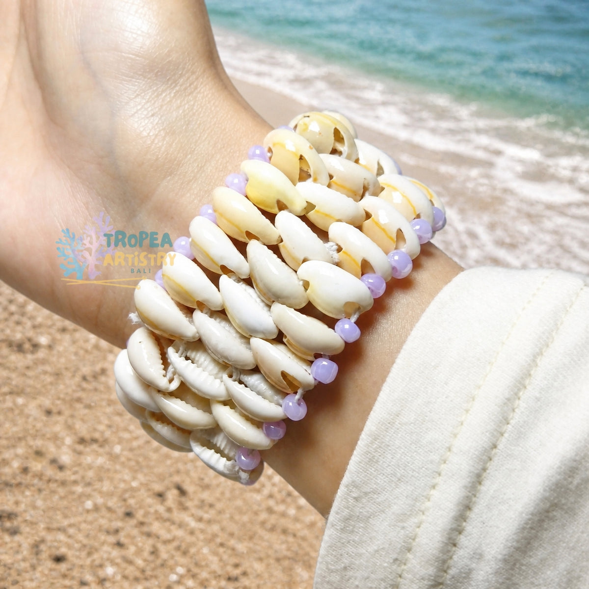 A close-up of a person's wrist wearing a multi-strand shell bracelet with small lavender beads, set against a beach background with sand and ocean water.