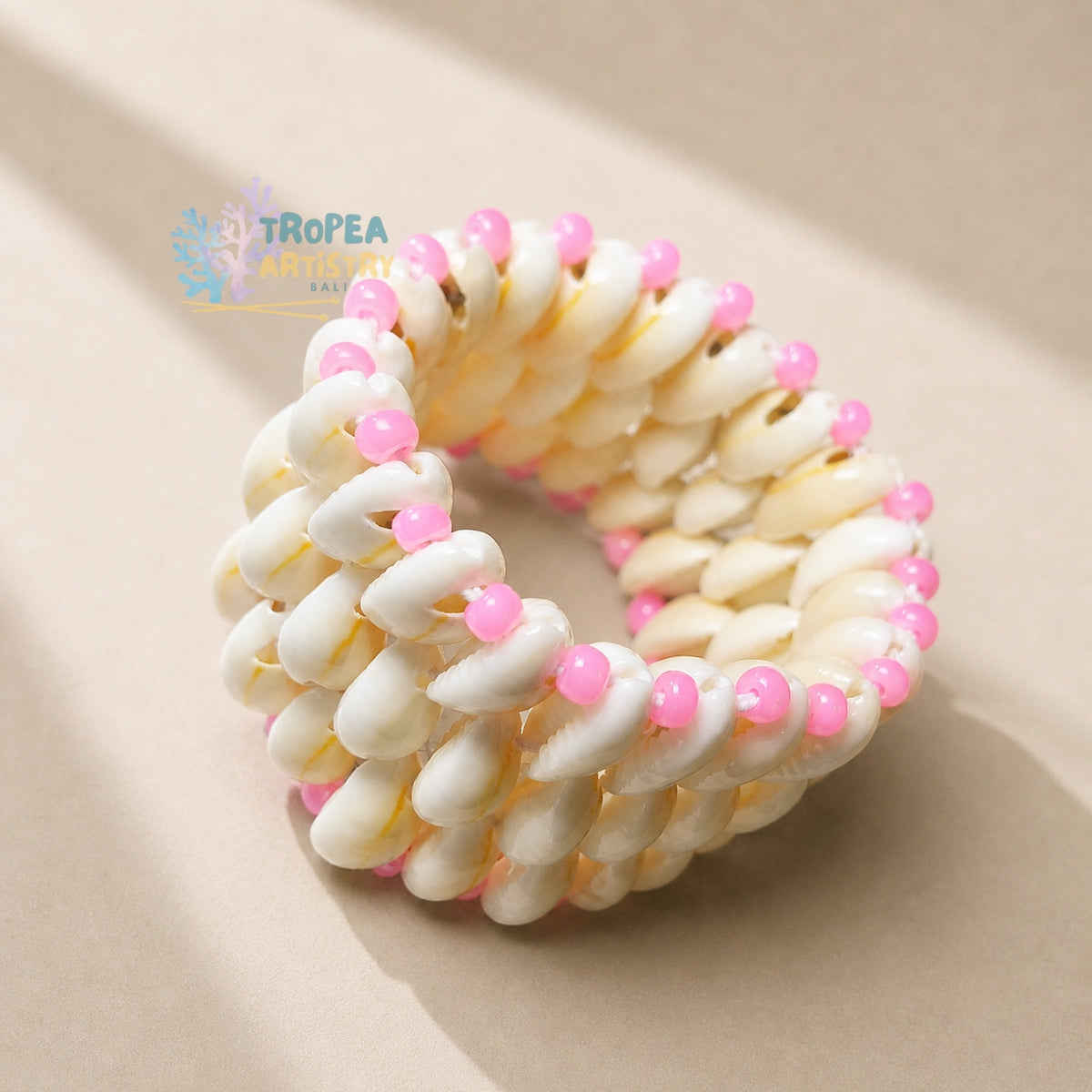 A woven shell bracelet made of cream-colored cowrie shells with small pink beads spaced along the outer edge, photographed on a warm beige background. A Tropea Aquarium watermark is visible in the upper left.