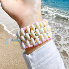 A close-up of a person's wrist wearing a multi-strand shell bracelet at the beach, with ocean waves and sand in the background. The bracelet is made of white cowrie shells with small pink beads along the edges.