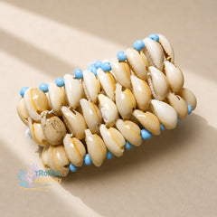 A handmade beaded bracelet made of cream-colored cowrie shells with light blue beads, displayed on a neutral beige background with soft shadows.