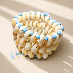 A coiled bracelet made of cream-colored cowrie shells with small light blue beads spaced along the front, photographed on a warm beige surface with soft shadows. A Trofea Artistry Italy watermark appears near the lower left.