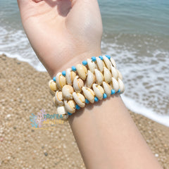 A close-up of a person's wrist wearing a multi-strand shell bracelet on a sandy beach with the ocean in the background. The bracelet features cream-colored cowrie shells with small bright blue beads separating them.