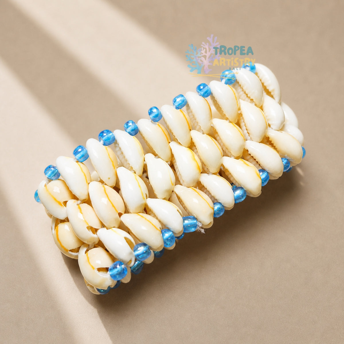 A woven bracelet made of white cowrie shells with small blue beads, displayed on a neutral beige background with soft shadows. A 'TROPEA' brand logo is visible in the upper right.