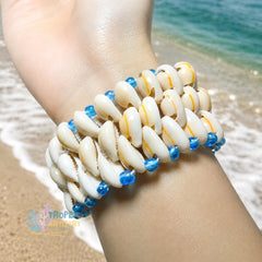 A close-up of a person's wrist wearing a woven bracelet made of white cowrie shells with small blue beads and thin yellow stripes, photographed on a sandy beach with ocean water in the background.
