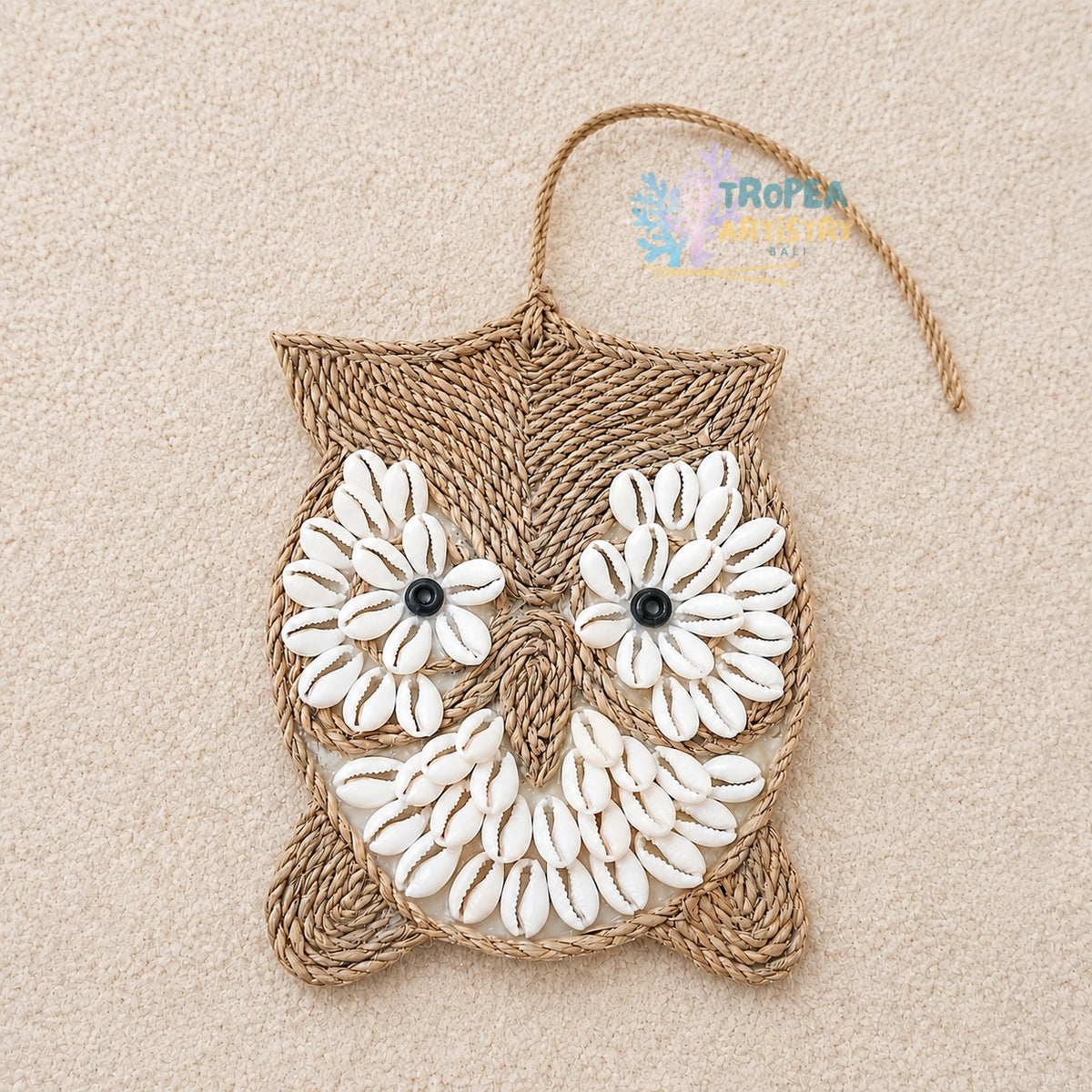 A handmade owl wall hanging crafted from natural jute rope and white cowrie shells, featuring black bead eyes and a hanging loop at the top, displayed on a textured beige background.