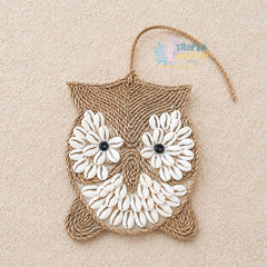 A handmade owl wall hanging crafted from natural jute rope and white cowrie shells, featuring black bead eyes and a hanging loop at the top, displayed on a textured beige background.
