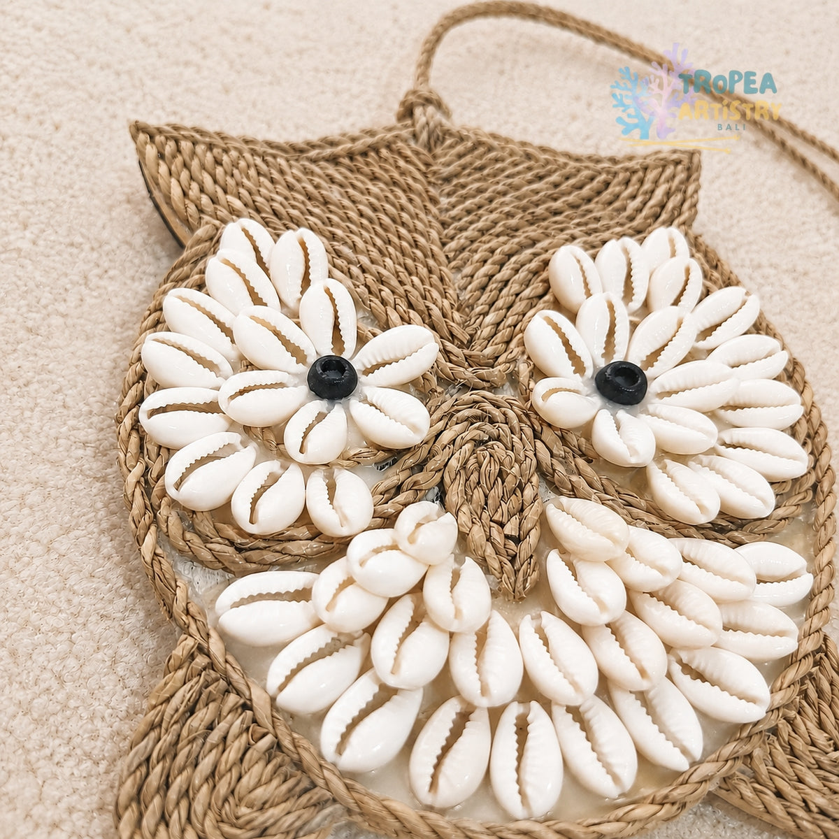 A woven raffia owl-shaped wall hanging or bag decorated with white cowrie shells forming flower eyes and a beak, with small black bead centers, shown on a beige textured background.