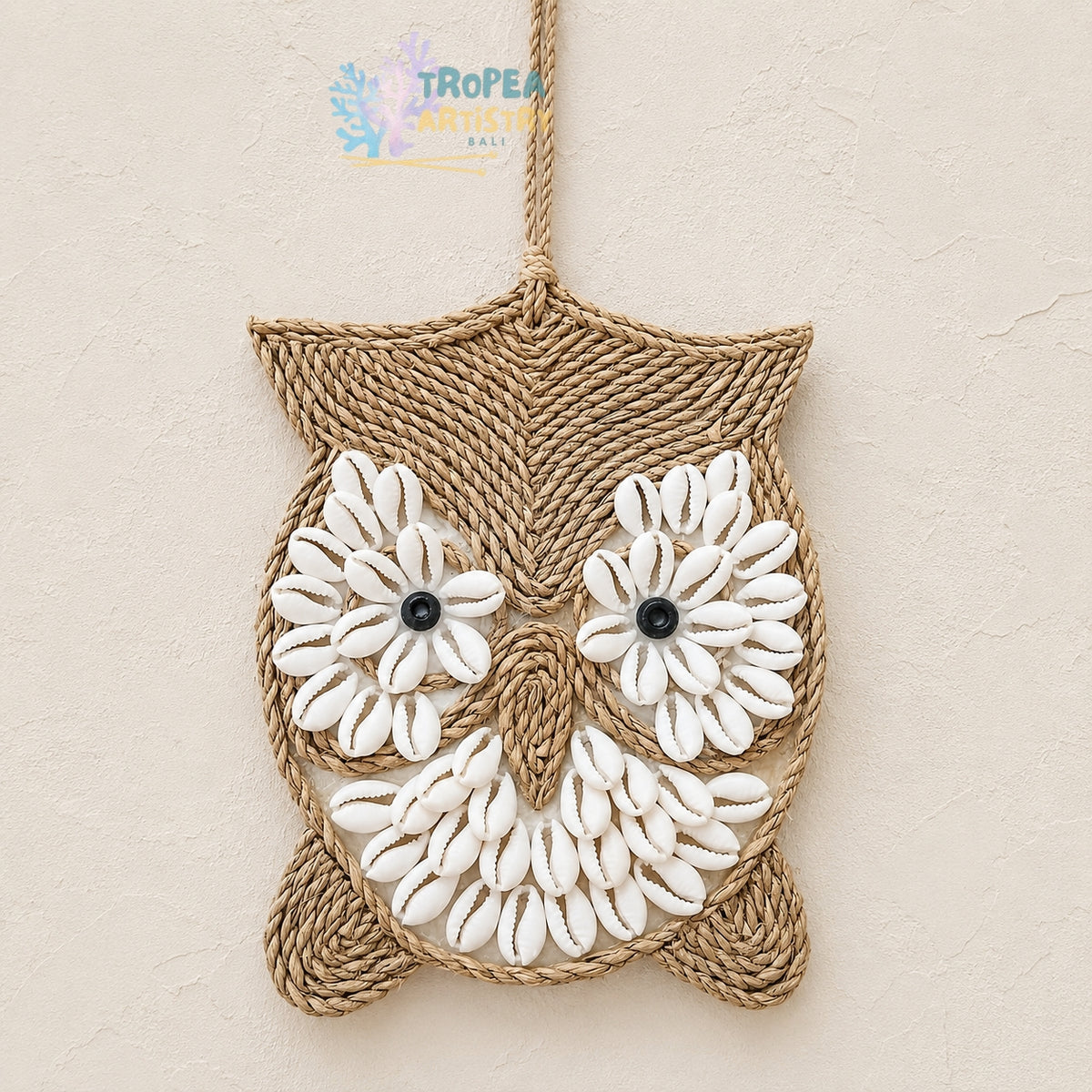 A handmade owl wall hanging made from natural jute rope and white cowrie shells, featuring black bead eyes, hanging against a textured beige wall.
