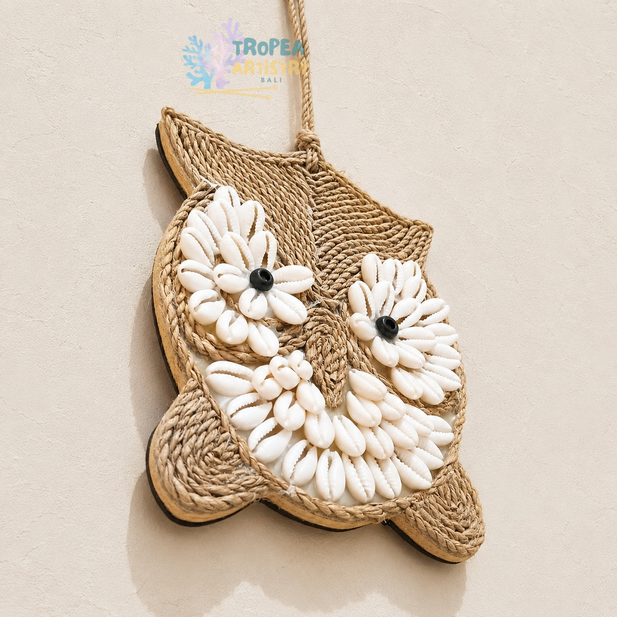 A pineapple-shaped wall hanging or ornament made from natural jute rope and white oval stones, photographed on a light beige textured background. A small 'TROPEA' watermark appears on the right side.
