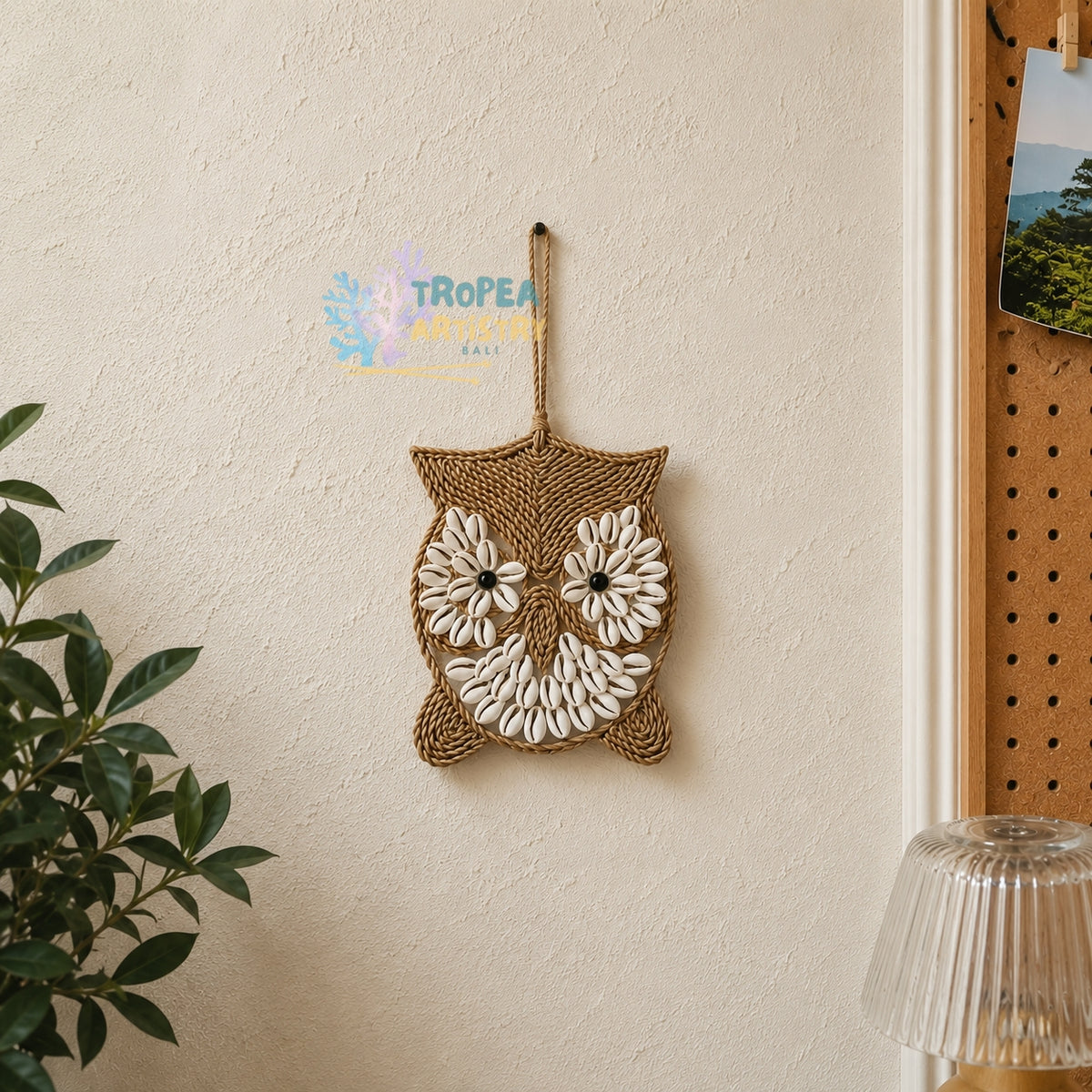 A woven owl wall hanging made from natural jute rope and white seashells, suspended by a rope loop on a textured beige wall. A green plant is visible on the left, and a wooden pegboard with a picture is on the right.