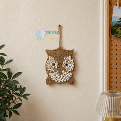 A woven owl wall hanging made from natural jute rope and white seashells, suspended by a rope loop on a textured beige wall. A green plant is visible on the left, and a wooden pegboard with a picture is on the right.