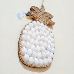 A pineapple-shaped decorative wall hanging made from natural woven raffia or straw, with white spherical beads covering the body, photographed on a plain light beige background.