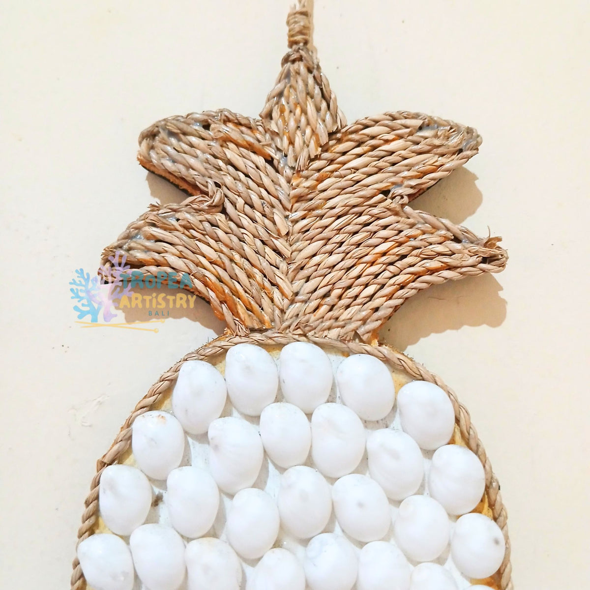A pineapple-shaped wall hanging made from woven natural fiber, with a leaf crown and a round body covered in white pom-pom beads, shown against a plain light beige background.
