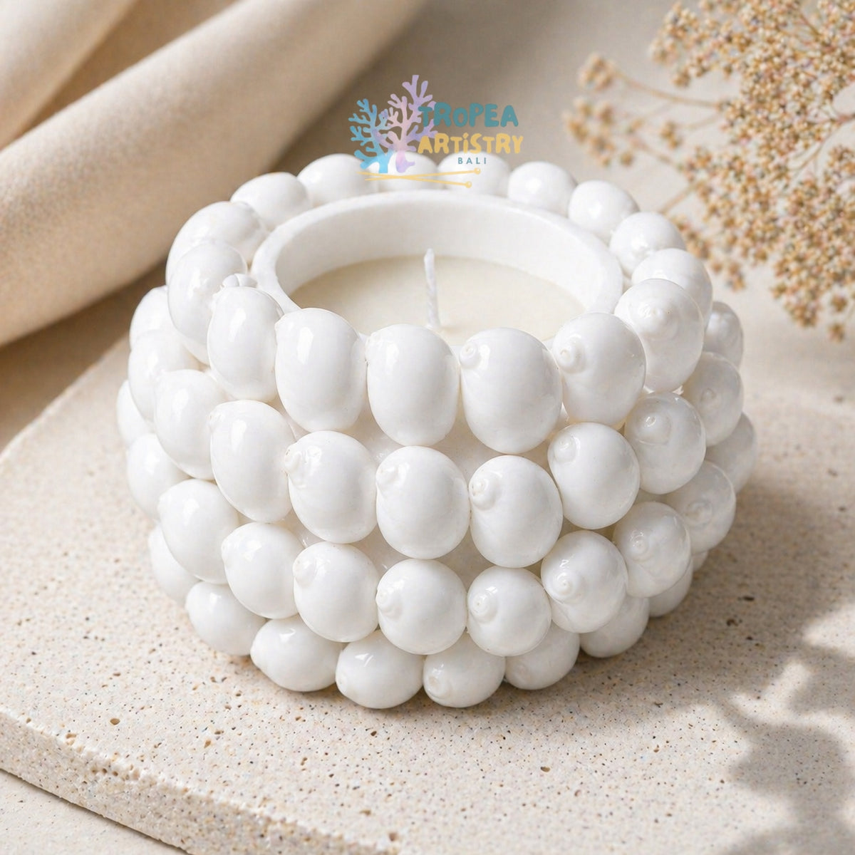 A white ceramic candle holder with a textured, beaded shell-like design, holding a white candle. The candle is styled on a light beige stone surface with neutral fabric and dried botanicals in the background.