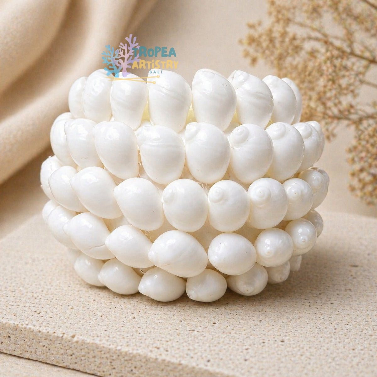 A white shell candle made of many small glossy shell-like pieces, photographed on a beige textured surface with soft neutral fabric and dried floral props in the background. A small 'Nopea Artistry' logo is visible near the top.