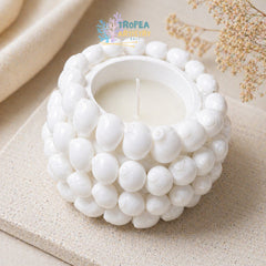 A white ceramic candle holder with a textured, beaded shell-like exterior, holding a white candle. The product is styled on a beige textured surface with neutral fabric and dried flowers in the background.