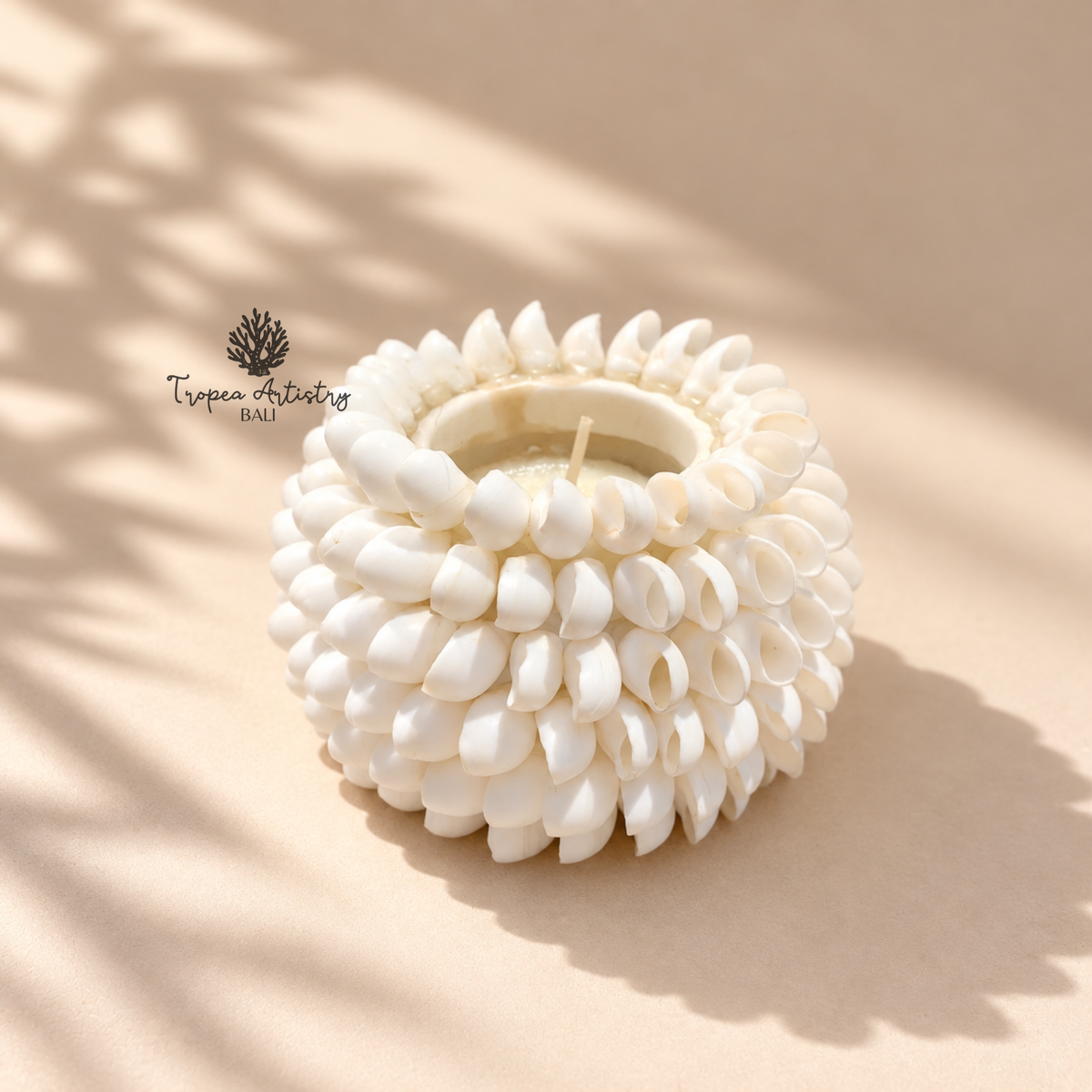 A white ceramic candle holder with a textured, flower-like petal design, containing a white wax candle, set against a warm beige background with soft leaf shadows.