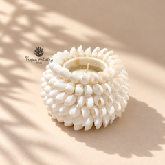 A white ceramic candle holder with a textured, flower-like petal design, containing a white wax candle, set against a warm beige background with soft leaf shadows.