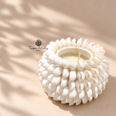 A white ceramic candle holder with a textured, flower-like layered design, containing a small white candle. The product is photographed on a warm beige surface with soft leaf shadows cast across it.