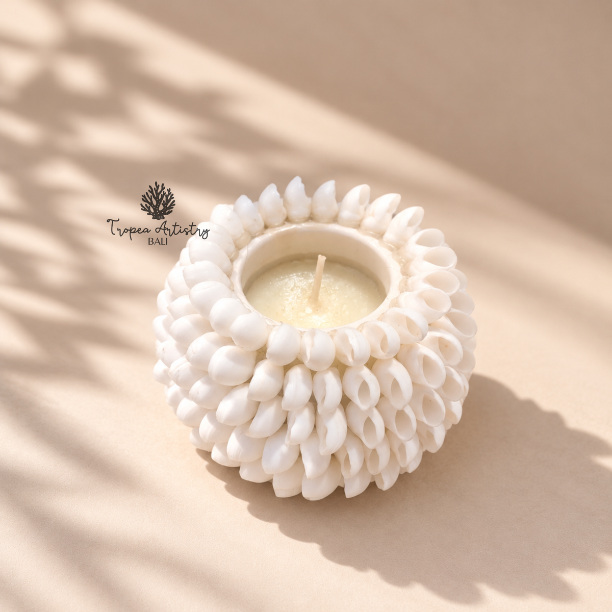 A white ceramic candle holder with a textured, shell-like floral pattern, holding a small cream-colored candle. The product is photographed on a warm beige surface with soft, artistic leaf shadows cast across it.