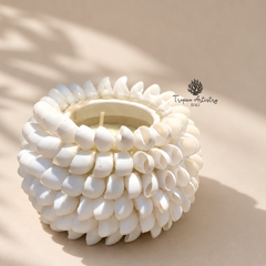 A white seashell candle holder with a textured, spiral shell pattern, containing a white wax candle. The product is photographed on a warm beige background with soft, natural shadows cast across it.