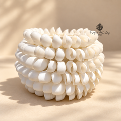 A white sculptural decorative object made of many rounded petal-like or shell-like pieces arranged in a rounded spiral stack, photographed on a warm beige surface with soft shadows. A small 'Tropic Arts Trading LLC' logo appears near the upper right.