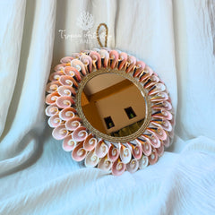 A round decorative wall mirror with a bohemian-style frame made of layered pink and white seashells, a natural wood center, and a rope-wrapped border. The mirror is hanging against draped white fabric, with a reflection of an interior room visible in the glass.