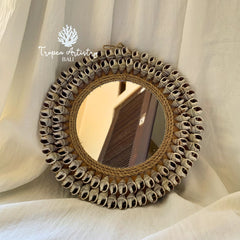A round decorative mirror with a hand-woven natural fiber frame and a border of small seashells, displayed against draped white fabric. The brand name 'Tropica Artistry Bali' is visible as an overlay in the upper left.
