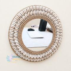 A round decorative wall mirror with a thick natural jute rope frame and a border of small white cowrie shells, mounted on a light beige wall. The reflection shows a desk setup with a keyboard and mouse.