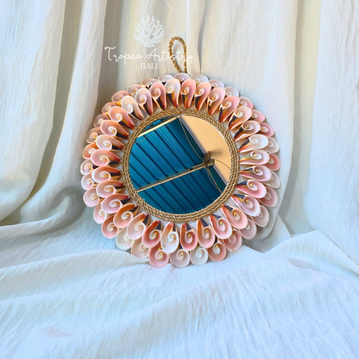 A decorative oval wall mirror with a nautical shell border and rope frame, hanging against draped white fabric. The shell pattern around the mirror is primarily pink, white, and beige, with a blue reflection visible in the mirror glass.