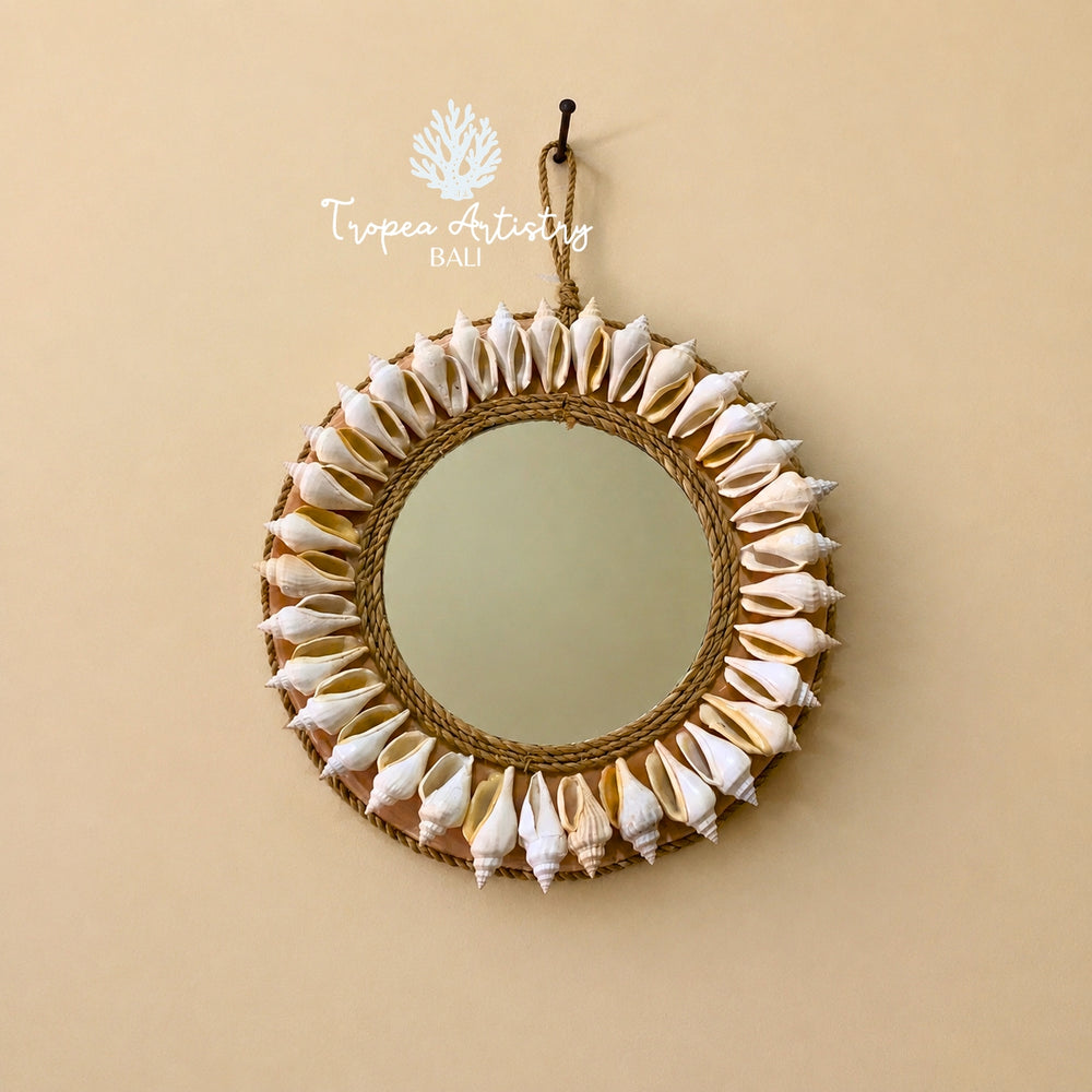 A round Bali-style wall mirror with a rope-wrapped frame and a border of white seashells, hanging from a hook on a beige wall. A 'Tropea Artistry Bali' logo is visible at the top.